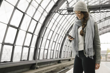 Glückliche junge Frau mit Smartphone auf dem Bahnsteig, Berlin, Deutschland - AHSF01425