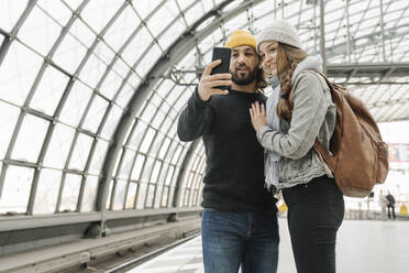 Glückliches junges Paar mit Smartphone auf dem Bahnsteig, Berlin, Deutschland - AHSF01436