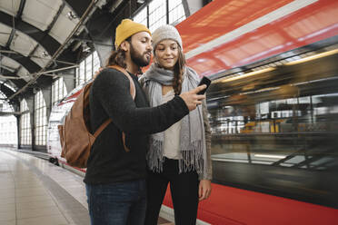 Junges Paar mit Smartphone auf dem Bahnsteig, während der Zug einfährt, Berlin, Deutschland - AHSF01452
