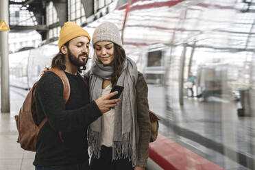 Junges Paar mit Smartphone auf dem Bahnsteig, während der Zug einfährt, Berlin, Deutschland - AHSF01500