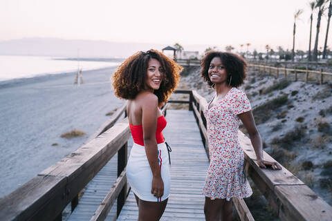 Junge lächelnde Frauen, die zusammen in der Nähe des Strandes spazieren gehen, sich umdrehen und in die Abendkamera schauen, lizenzfreies Stockfoto