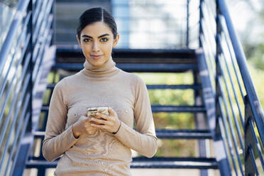 Porträt einer selbstbewussten jungen Frau mit Smartphone auf einer Außentreppe - JSMF01366