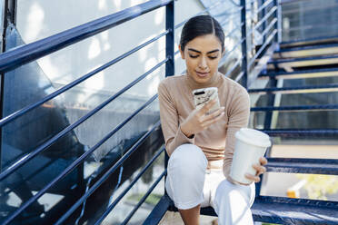 Junge Frau mit Getränk zum Mitnehmen sitzt auf einer Außentreppe und benutzt ihr Smartphone - JSMF01369