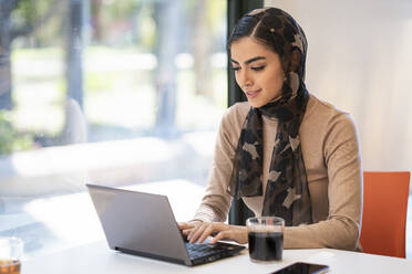 Young woman wearing headscarf using laptop in a cafe - JSMF01382