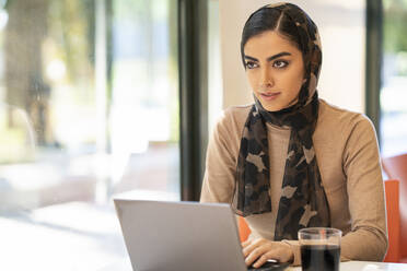 Young woman wearing headscarf using laptop in a cafe - JSMF01383