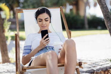 Junge Frau sitzt in einem Liegestuhl, trägt Kopfhörer und benutzt ein Smartphone - JSMF01384