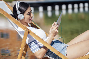 Junge Frau sitzt in einem Liegestuhl, trägt Kopfhörer und benutzt ein Tablet - JSMF01387