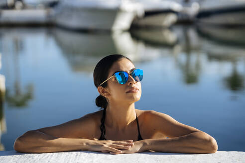 Porträt einer jungen Frau mit Sonnenbrille und Bikini an einer Kaimauer lehnend - JSMF01413