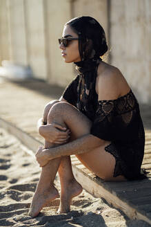 Junge Frau mit Kopftuch und Sonnenbrille am Strand sitzend - JSMF01426
