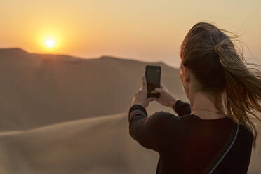 Frau macht ein Smartphone-Foto in der Wüste bei Sonnenuntergang, Düne 7, Walvis Bay, Namibia - VEGF00958