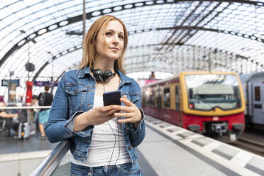 Lächelnde Frau mit Smartphone und Kopfhörern auf dem Bahnsteig, Berlin, Deutschland - WPEF02321