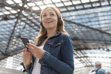 Lächelnde Frau mit Smartphone und Kopfhörern am Bahnhof, Berlin, Deutschland - WPEF02324