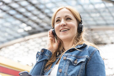 Glückliche Frau, die mit Kopfhörern am Bahnhof Musik hört, Berlin, Deutschland - WPEF02325