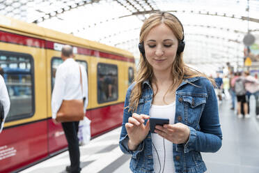 Smiling woman with smartphone and headphones on the station platform, Berlin, Germany - WPEF02326