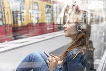 Frau mit Smartphone und Kopfhörern wartet am Bahnhof, Berlin, Deutschland - WPEF02330