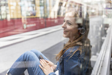 Glückliche Frau mit Smartphone und Kopfhörern beim Warten am Bahnhof, Berlin, Deutschland - WPEF02331