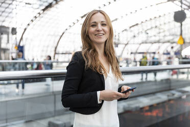 Porträt einer lächelnden Frau mit Smartphone am Bahnhof, Berlin, Deutschland - WPEF02335
