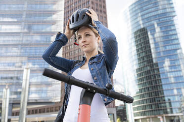 Woman with e-scooter in the city putting on helmet, Berlin, Germany - WPEF02344