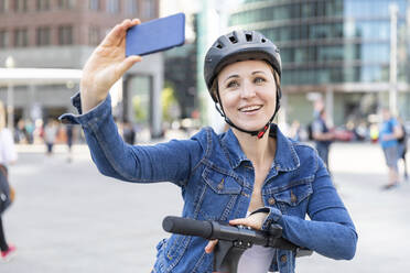 Lächelnde Frau mit E-Scooter, die ein Selfie in der Stadt macht, Berlin, Deutschland - WPEF02352