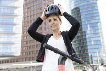 Woman with e-scooter in the city putting on helmet, Berlin, Germany - WPEF02353