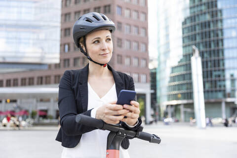 Frau beim Pendeln mit E-Scooter und Smartphone in der Stadt, Berlin, Deutschland, lizenzfreies Stockfoto