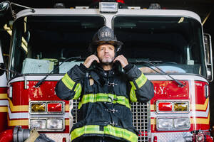 USA, New York, Porträt eines Feuerwehrmanns, der seinen Helm vor einem Feuerwehrauto aufsetzt - CJMF00184
