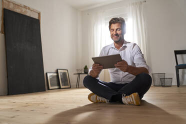 Porträt eines lächelnden reifen Mannes, der zu Hause auf dem Boden sitzt und ein digitales Tablet benutzt - PHDF00020