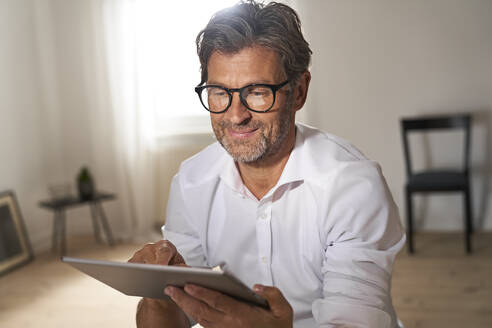 Porträt eines lächelnden reifen Mannes mit Brille, der zu Hause ein digitales Tablet benutzt - PHDF00022
