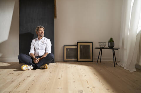 Portrait of relaxed mature man sitting on the floor at home looking at distance stock photo