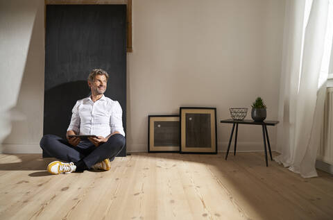 Entspannter reifer Mann mit digitalem Tablet, der zu Hause auf dem Boden sitzt und in die Ferne schaut, lizenzfreies Stockfoto