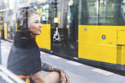 Porträt einer tätowierten jungen Frau, die an einer Straßenbahnhaltestelle wartet, Berlin, Deutschland - WPEF02369