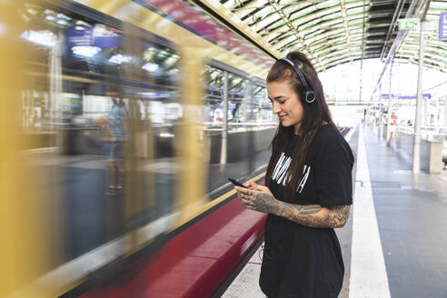 Tätowierte junge Frau mit Kopfhörern steht am Bahnsteig und benutzt ihr Smartphone, Berlin, Deutschland - WPEF02373