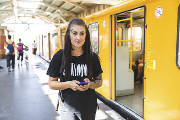 Porträt einer tätowierten jungen Frau mit Smartphone am Bahnsteig, Berlin, Deutschland - WPEF02381