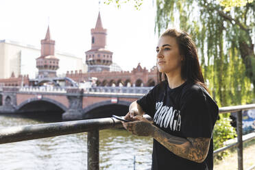 Porträt einer tätowierten jungen Frau mit Smartphone an der Spree, Berlin, Deutschland - WPEF02383