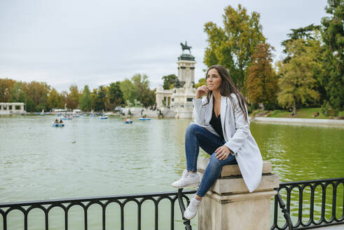 Nachdenkliche Frau sitzt in der Nähe eines Sees im El Retiro Park, Madrid, Spanien - KIJF02838