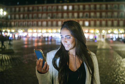 Frau benutzt ihr Smartphone auf der Plaza Mayor bei Nacht, Madrid, Spanien - KIJF02847