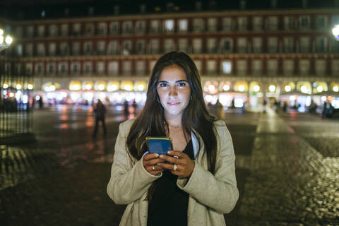 Porträt einer Frau, die ihr Smartphone auf der Plaza Mayor bei Nacht benutzt, Madrid, Spanien, lizenzfreies Stockfoto