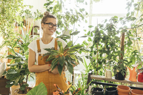 Glückliche junge Frau hält eine Pflanze in einer kleinen Gärtnerei, lizenzfreies Stockfoto