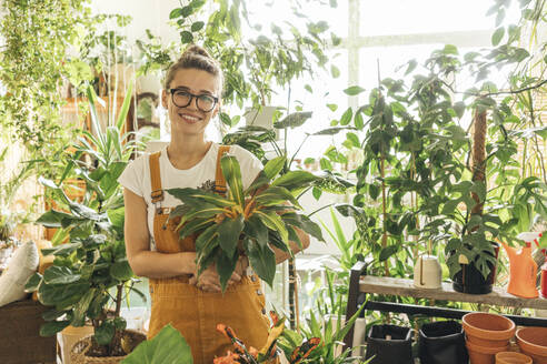 Porträt einer lächelnden jungen Frau, die eine Pflanze in einer kleinen Gärtnerei hält - VPIF01841
