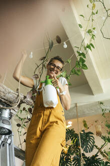 Happy young woman standing on a ladder caring for plants in a small shop - VPIF01843