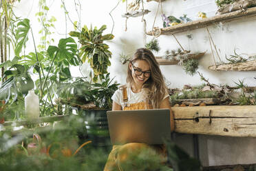 Happy young woman using laptop in a small gardening shop - VPIF01852