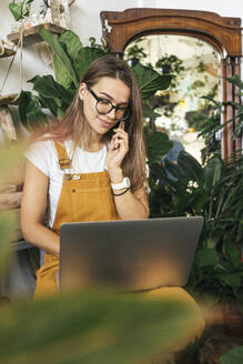 Young woman with laptop on the phone in a small gardening shop - VPIF01859