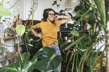 Fröhliche junge Frau umgeben von Pflanzen, die mit Kopfhörern Musik hört - VPIF01877