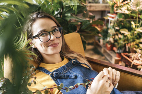 Portrait of a smiling young woman sitting in armchair surrounded by plants - VPIF01881