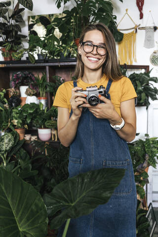 Porträt einer glücklichen jungen Frau mit einer Kamera in einer kleinen Gärtnerei, lizenzfreies Stockfoto