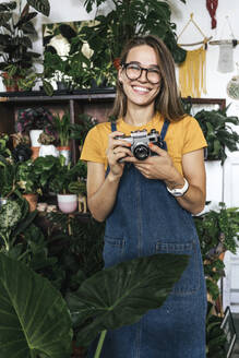 Porträt einer glücklichen jungen Frau mit einer Kamera in einer kleinen Gärtnerei - VPIF01889
