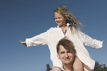 Vater trägt seine kleine Tochter auf den Schultern unter blauem Himmel - EYAF00722