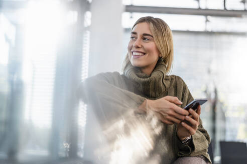 Smiling young woman with cell phone sitting in waiting area looking around - DIGF08967