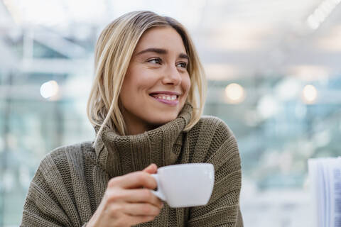 Porträt einer lächelnden jungen Frau, die eine Tasse Kaffee hält und wegschaut, lizenzfreies Stockfoto