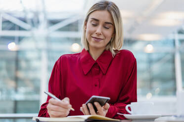 Junge Geschäftsfrau mit Handy macht sich Notizen in einem Café - DIGF08978